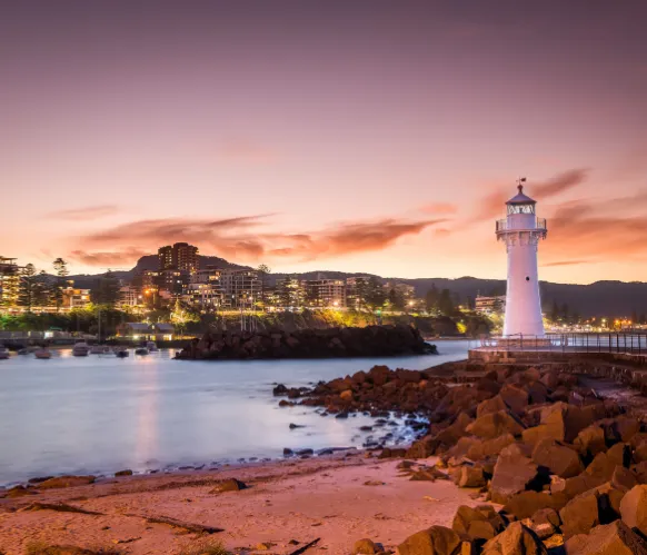 Wollongong Harbour lighthouse at sunset with city lights and rocky shoreline in the foreground.